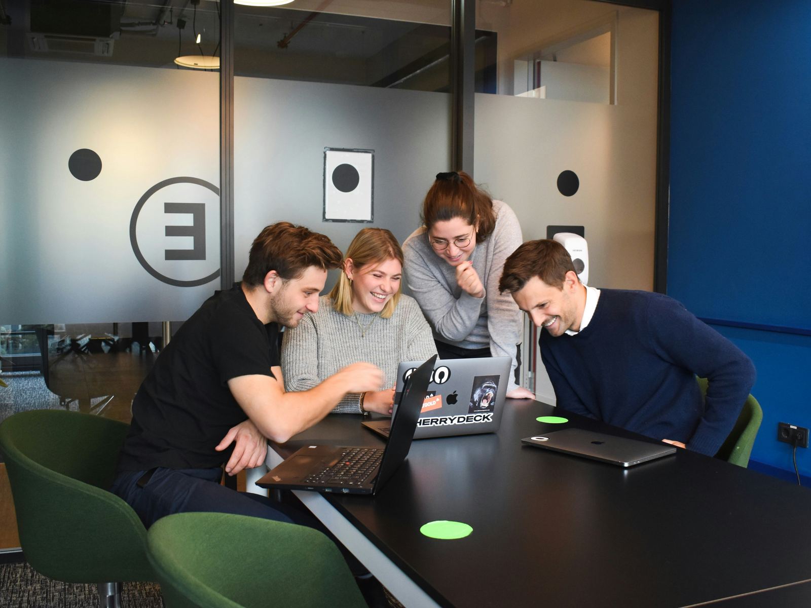 Four coworkers gathered closely around a laptop, laughing together, the kind of shared attention 'The Culture Code' calls a belonging cue