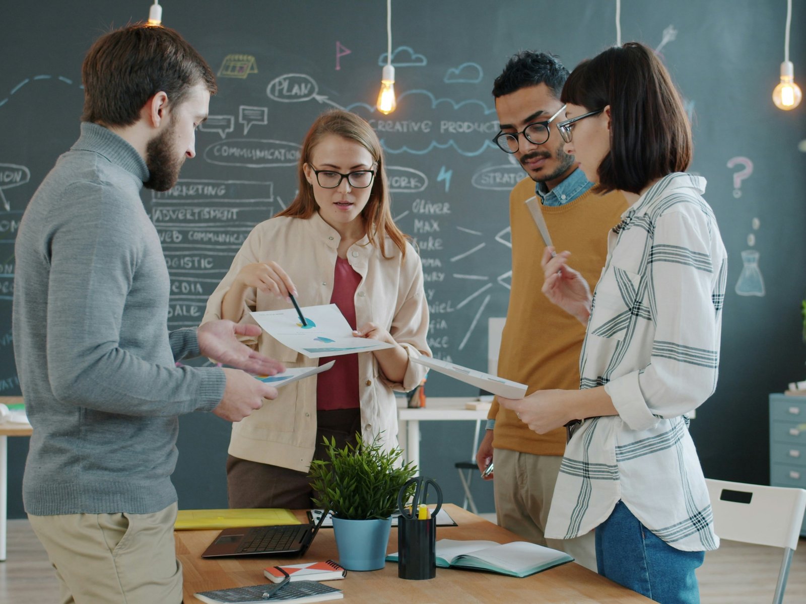Four coworkers standing around a table reviewing printed charts together in front of a chalkboard wall covered in business planning diagrams