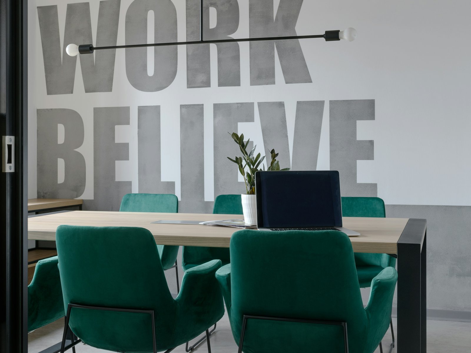 An empty conference room with the words "Love, Work, Believe" painted on the wall behind an unused meeting table, a laptop left open with no one there