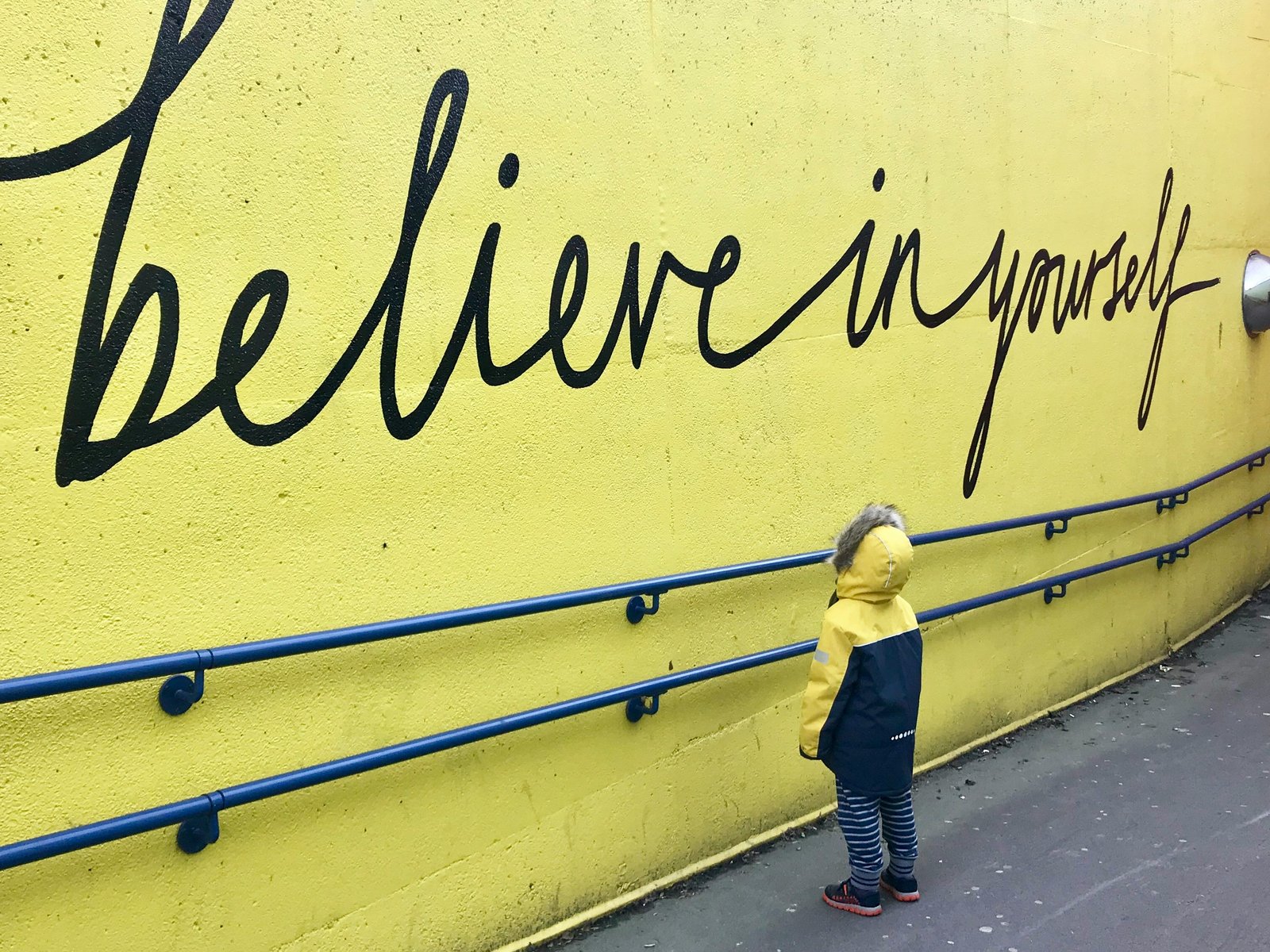 A small figure standing alone at the base of a wall painted with the words "believe in yourself," dwarfed by the empty space around them