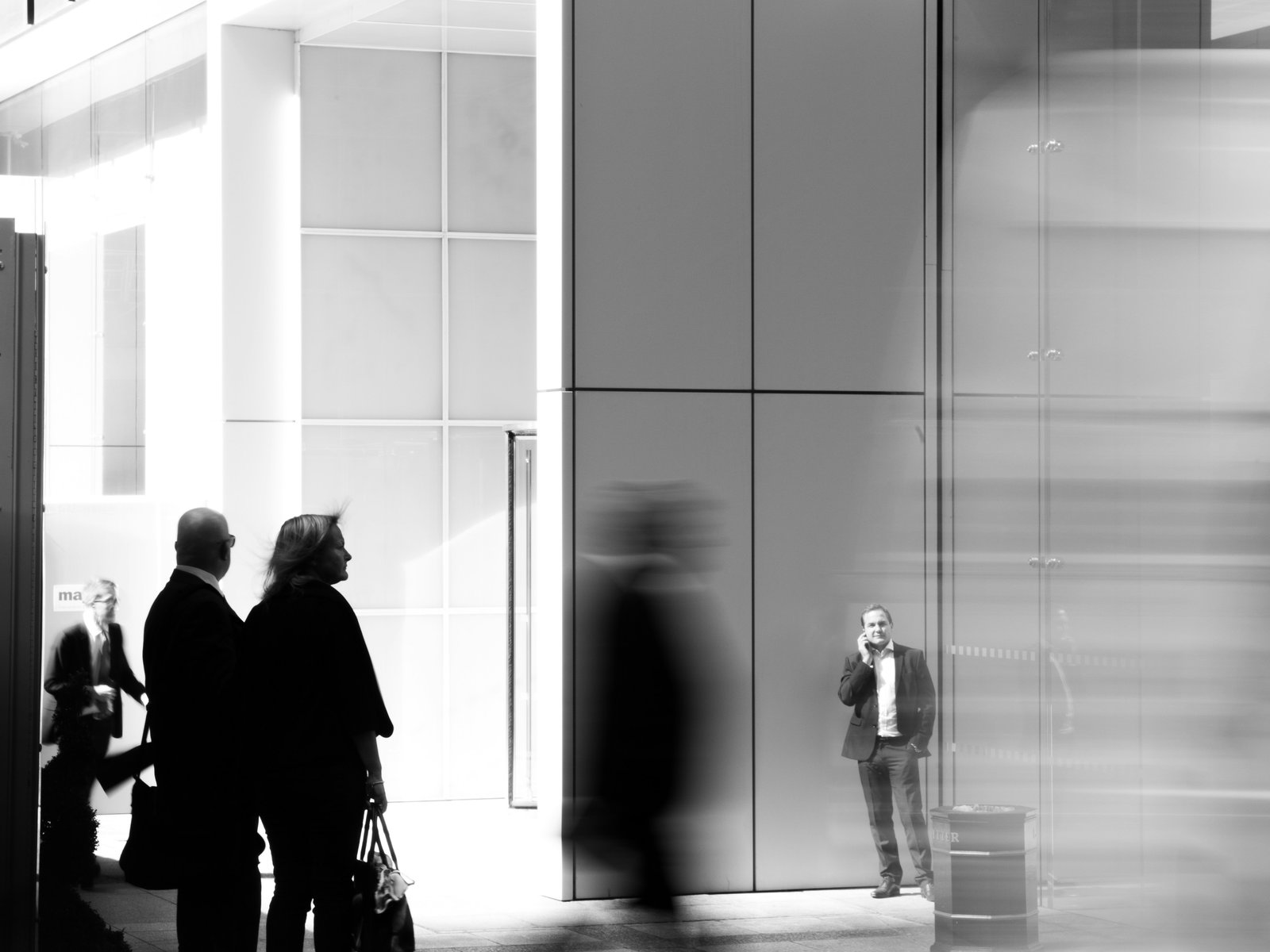 A blurred pedestrian walks past a still businessman on a phone call outside a glass office building in black and white, a picture of the manager who stays on the job while the support around them moves on