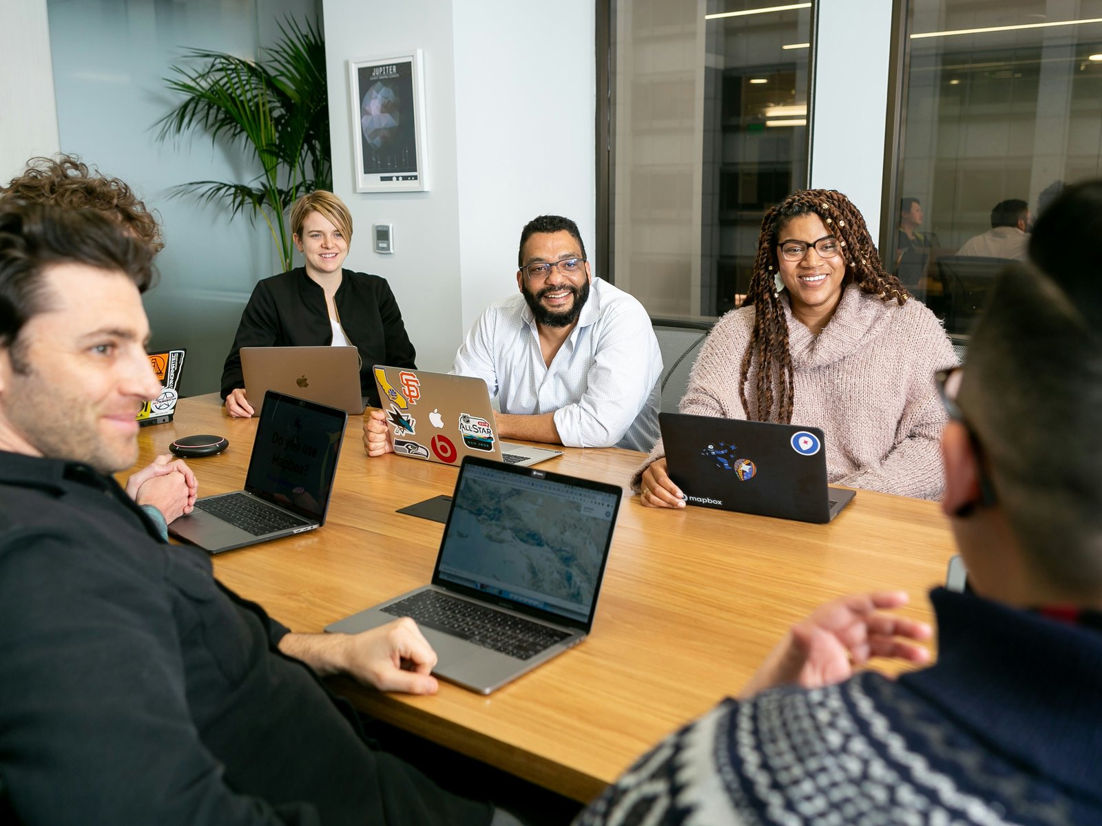 Five coworkers seated around a wooden conference table with open laptops, smiling and listening as a colleague speaks during a team meeting