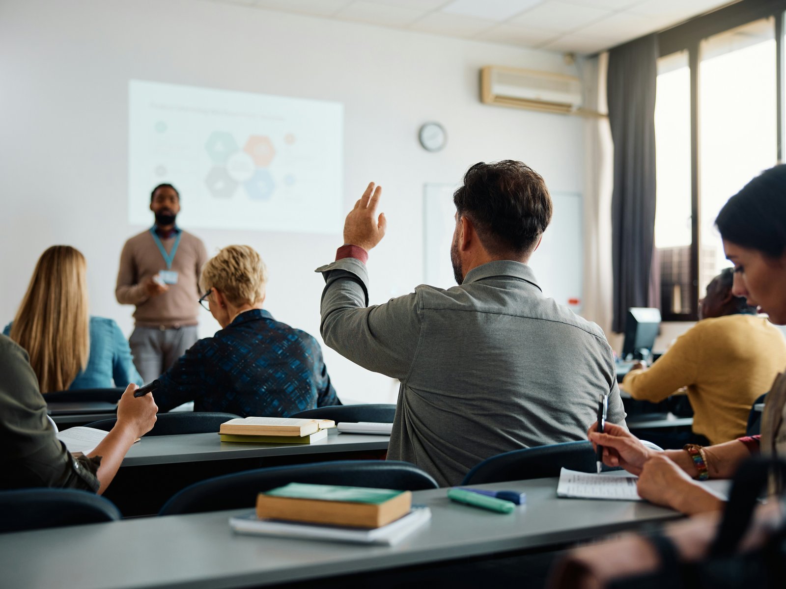 A man in a gray shirt raising his hand to ask a question during a workplace training session while a presenter stands near a projected diagram, an image of employee development in action